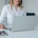 Consultor de Atendimento Home Office e mais 16 Vagas A woman working at a desk using a laptop and smartphone, exemplifying remote work.