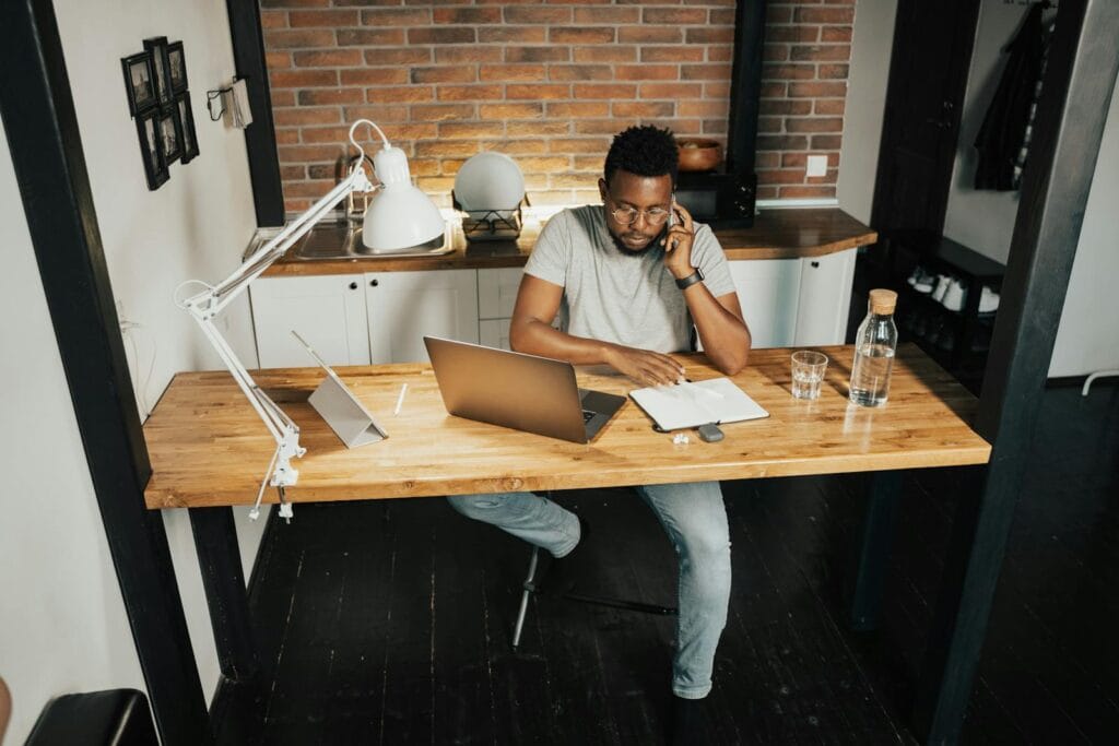 44 Vagas de Emprego na Comm Center A young man at a desk in a home office, using a laptop and phone, embodying remote work lifestyle.