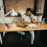 44 Vagas de Emprego na Comm Center A young man at a desk in a home office, using a laptop and phone, embodying remote work lifestyle.