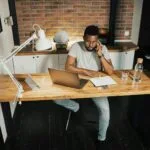 44 Vagas de Emprego na Comm Center A young man at a desk in a home office, using a laptop and phone, embodying remote work lifestyle.