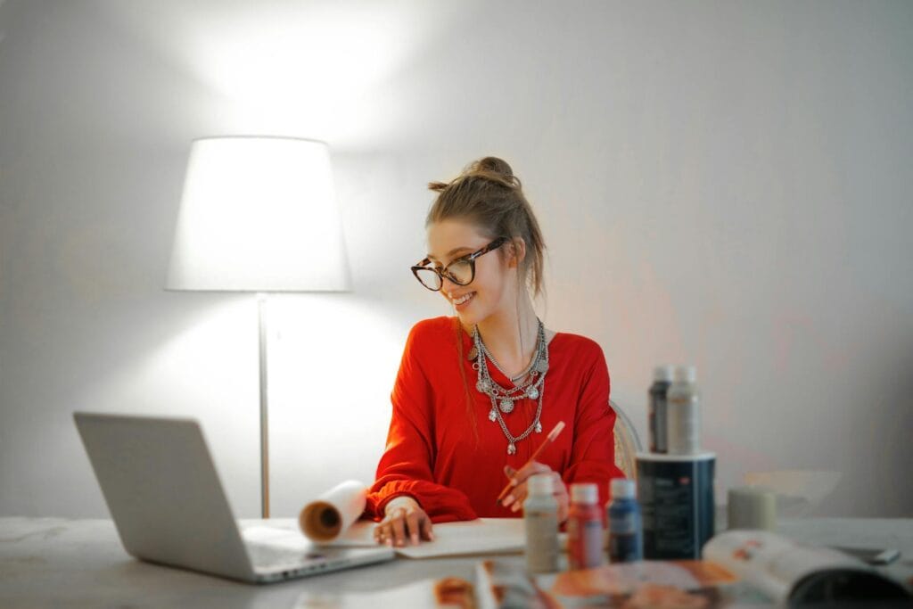 150 Vagas Remotas para quem tem o Ensino Médio! Smiling woman in red using laptop and art supplies at home office desk.