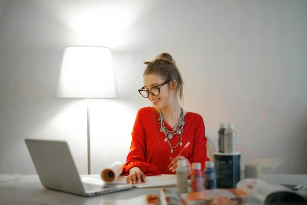 150 Vagas Remotas para quem tem o Ensino Médio! Smiling woman in red using laptop and art supplies at home office desk.