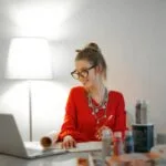 150 Vagas Remotas para quem tem o Ensino Médio! Smiling woman in red using laptop and art supplies at home office desk.